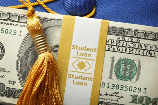 A stack of one hundred dollar bills in a money wrapper labeled "Student Loan" on top of a blue graduation cap. A gold graduation tassel is draped over the stack of money.